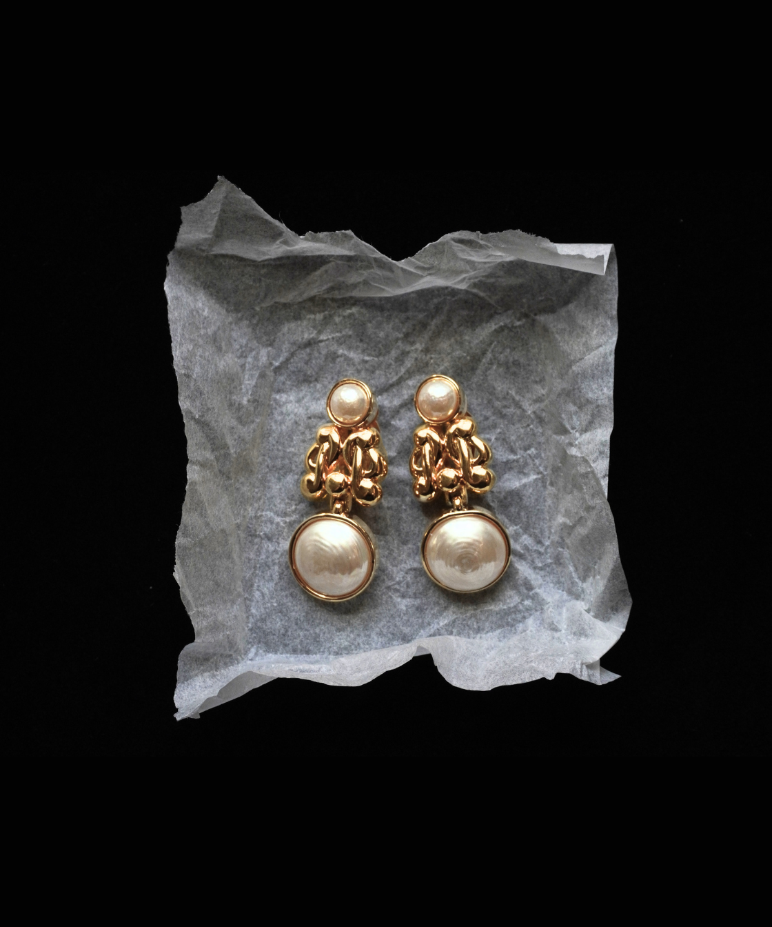 A pair of gold-toned drop earrings with textured floral clusters and large round pearl-like drops, displayed on a crumpled translucent sheet against a black background.
