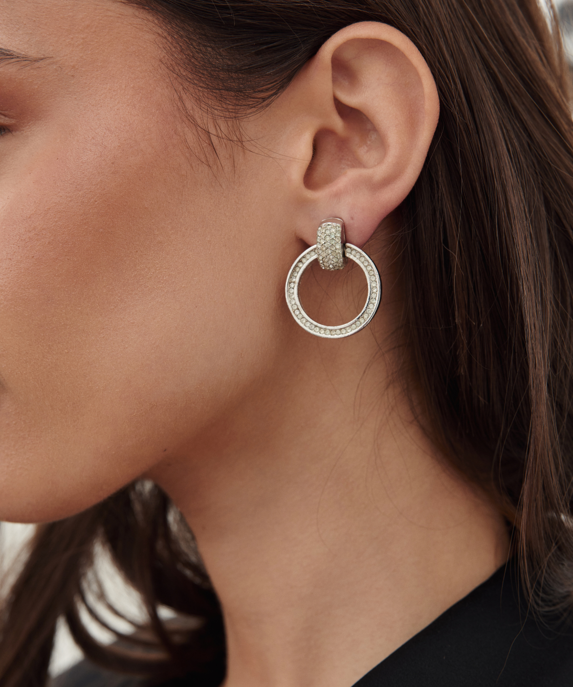A close-up of a woman's ear wearing a silver-toned double-hoop earring adorned with small clear stones.