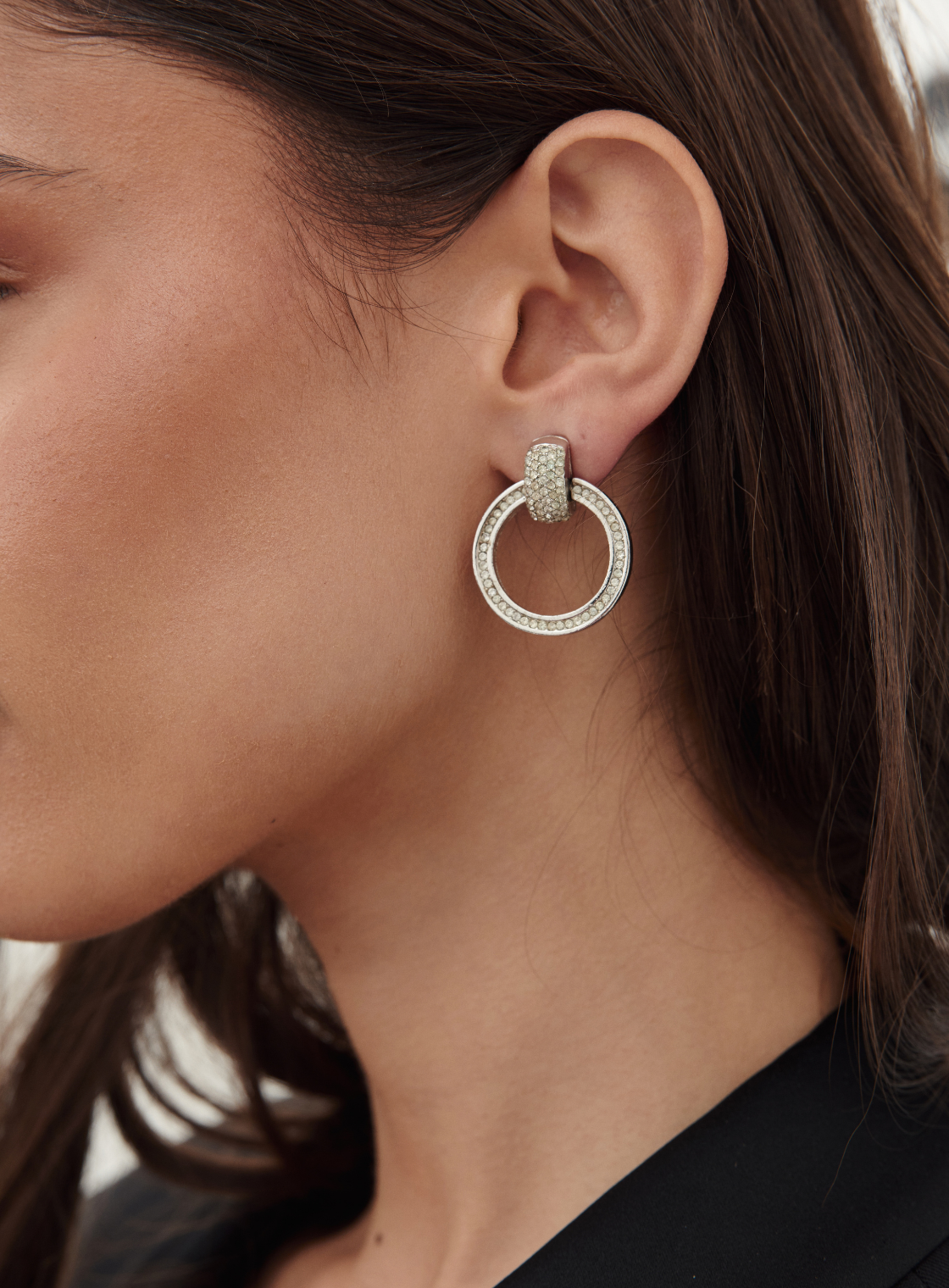 A close-up of a woman's ear wearing a silver-toned double-hoop earring adorned with small clear stones.