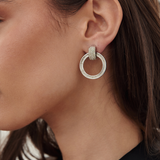 A close-up of a woman's ear wearing a silver-toned double-hoop earring adorned with small clear stones.