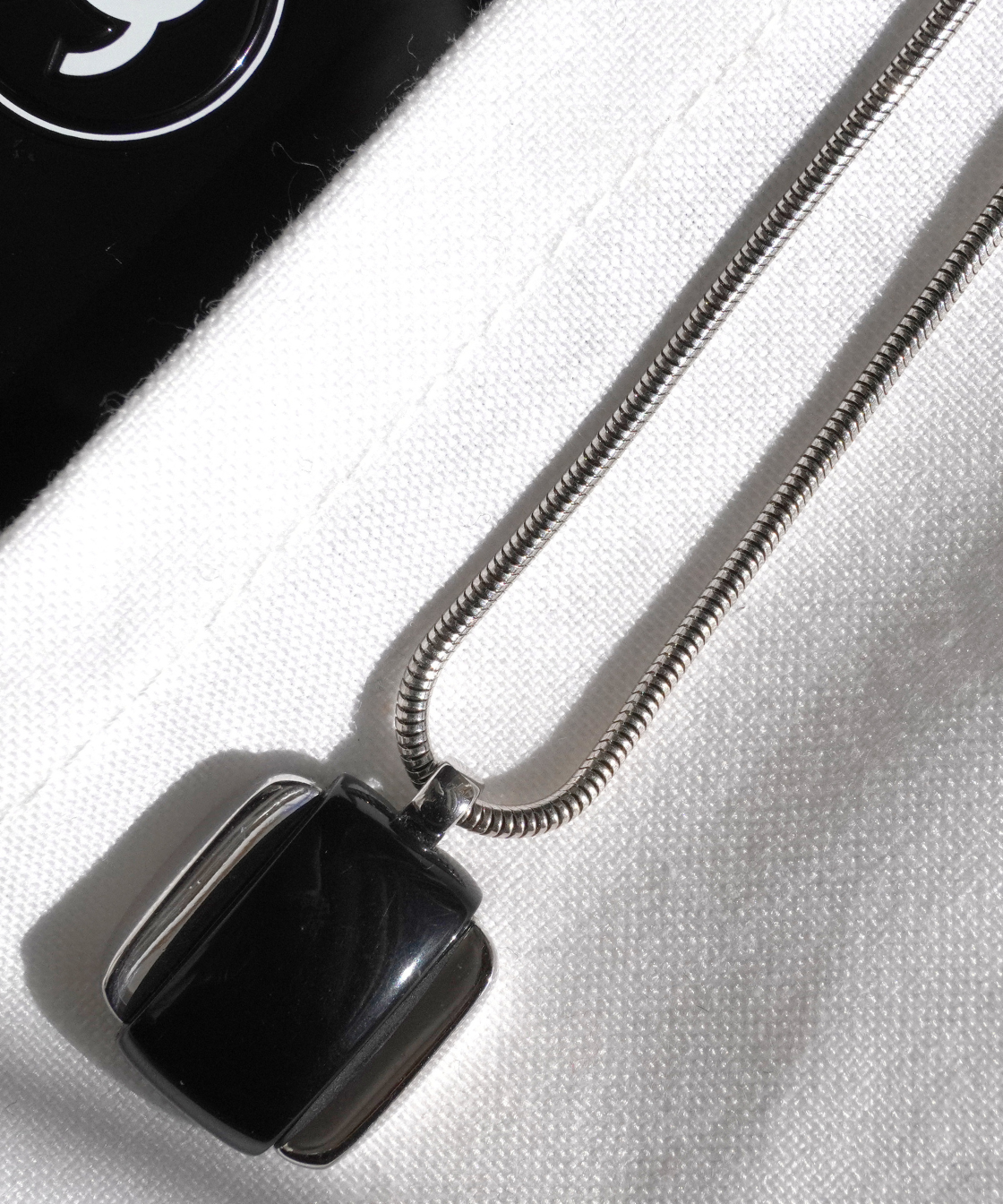 A close-up of a silver snake chain Givenchy necklace with a black square pendant, resting on a white textured fabric next to a black surface with a white Chanel logo.
