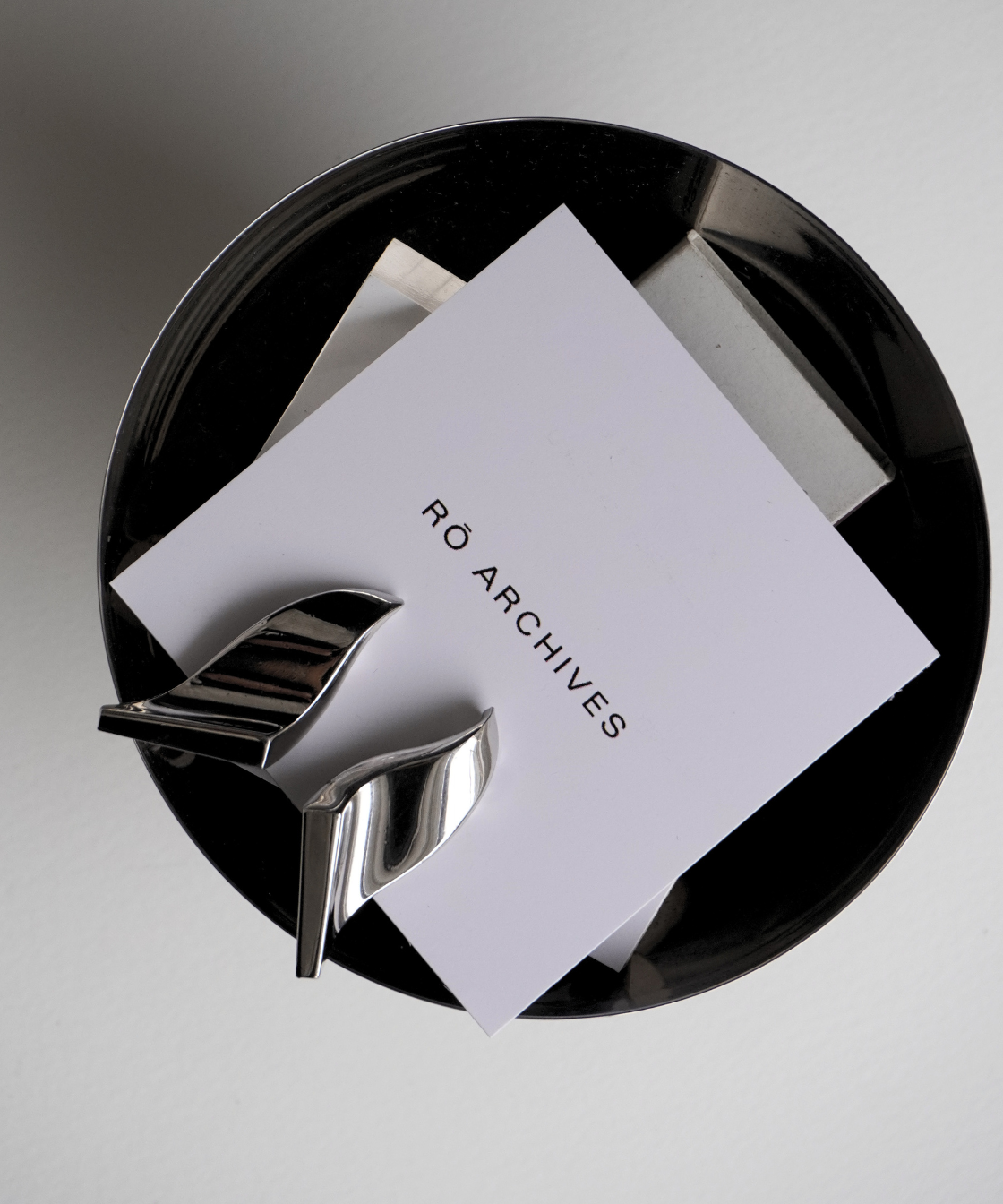 A white square card printed with the text 'RØ ARCHIVES' in black, held in a reflective silver metal clip Errings inside a glossy black circular tray on a light gray background.