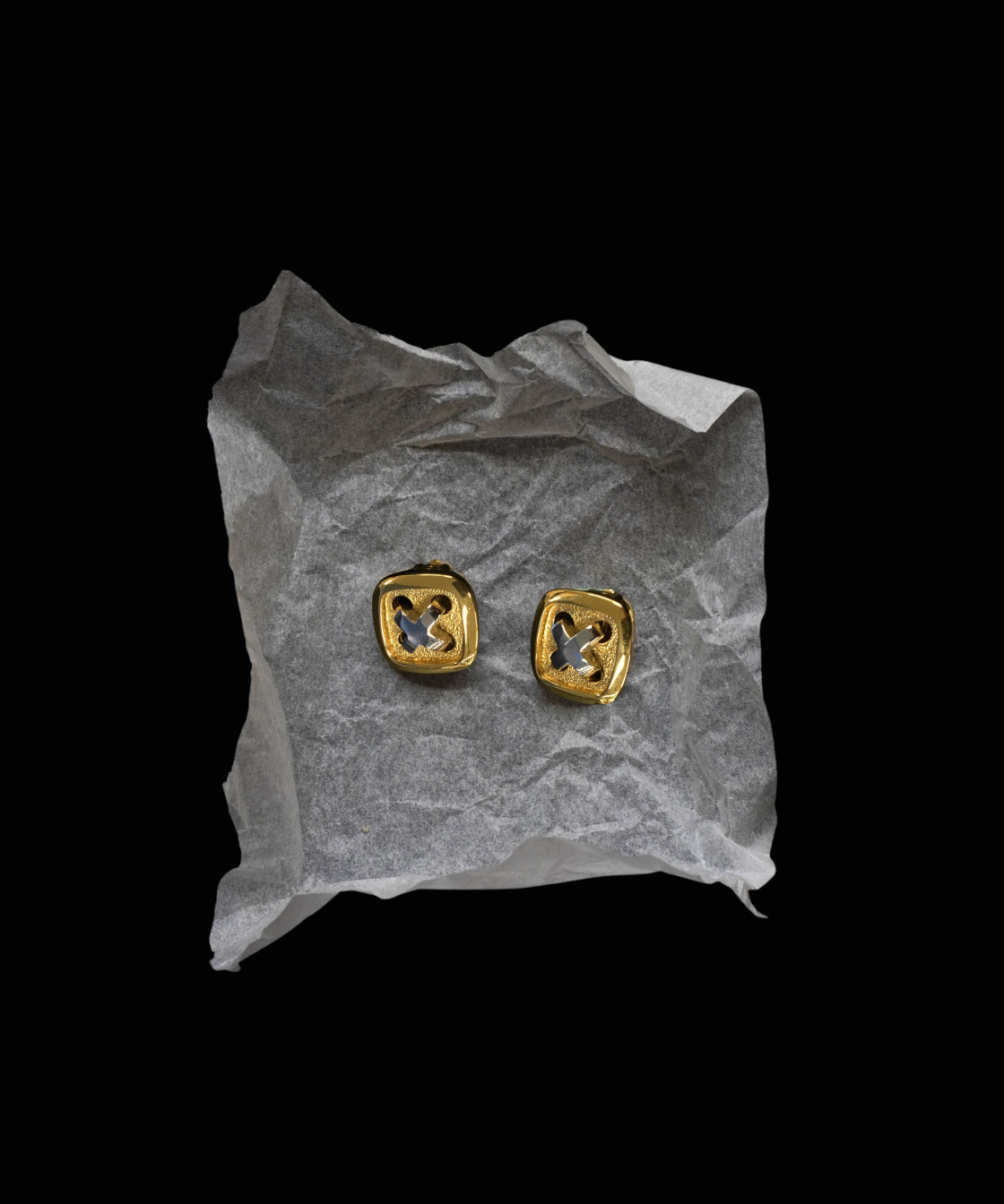 A pair of gold square stud earrings with a cutout geometric design, displayed on a crumpled piece of white tissue paper against a solid black background.
