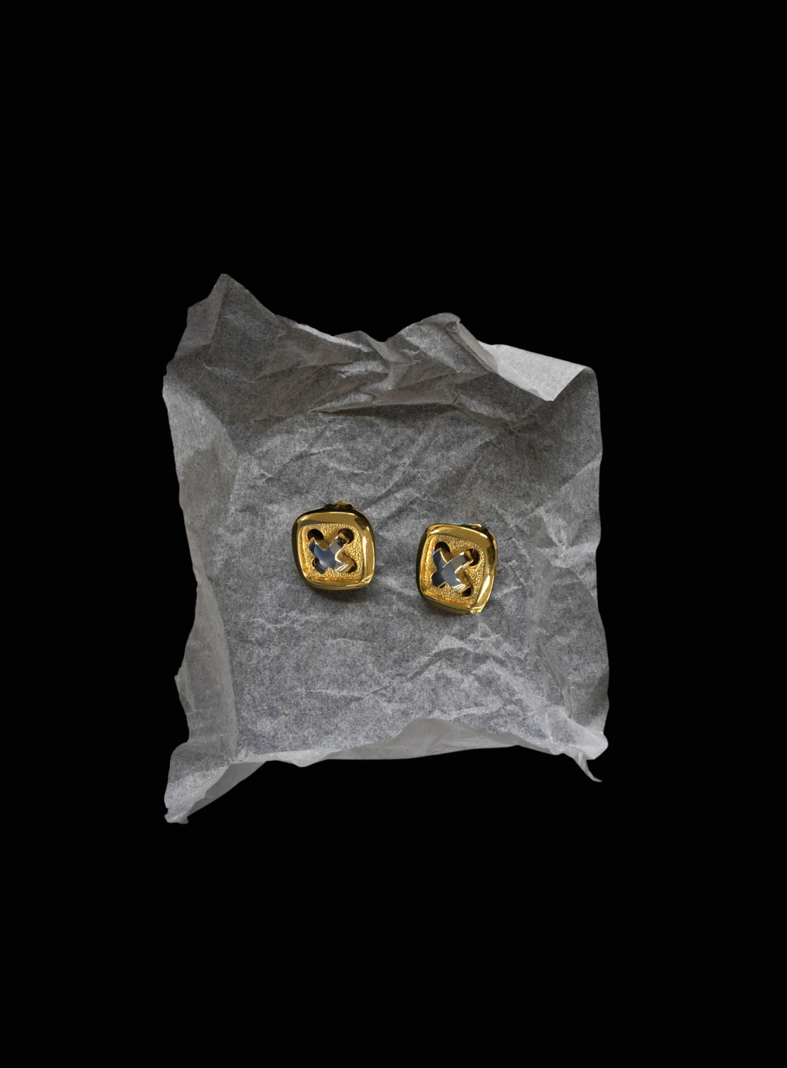 A pair of gold square stud earrings with a cutout geometric design, displayed on a crumpled piece of white tissue paper against a solid black background.
