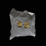 A pair of gold square stud earrings with a cutout geometric design, displayed on a crumpled piece of white tissue paper against a solid black background.