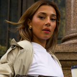 A woman wearing a beige trench coat over a white t-shirt, holding a brown and black Fendi monogram handbag, with a coffee cup in her hand. She is wearing a gold earring.