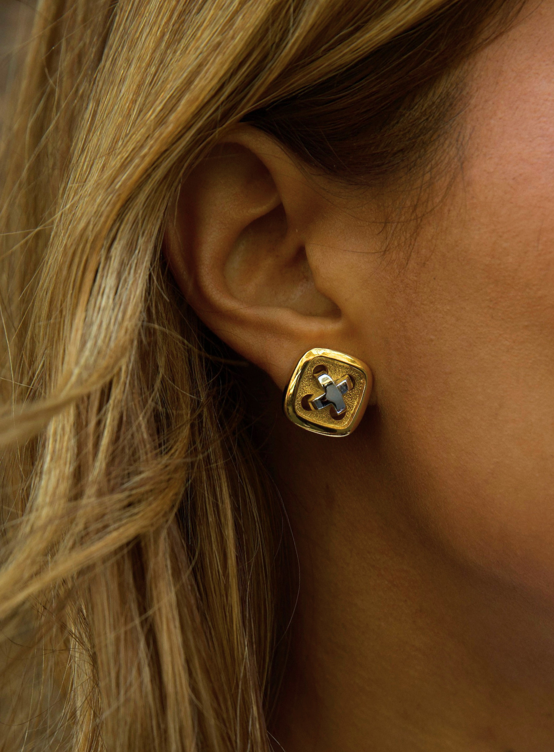 A close-up of a person's ear wearing a gold square stud earring with a blue floral or starburst motif in the center.