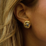 A close-up of a person's ear wearing a gold square stud earring with a blue floral or starburst motif in the center.