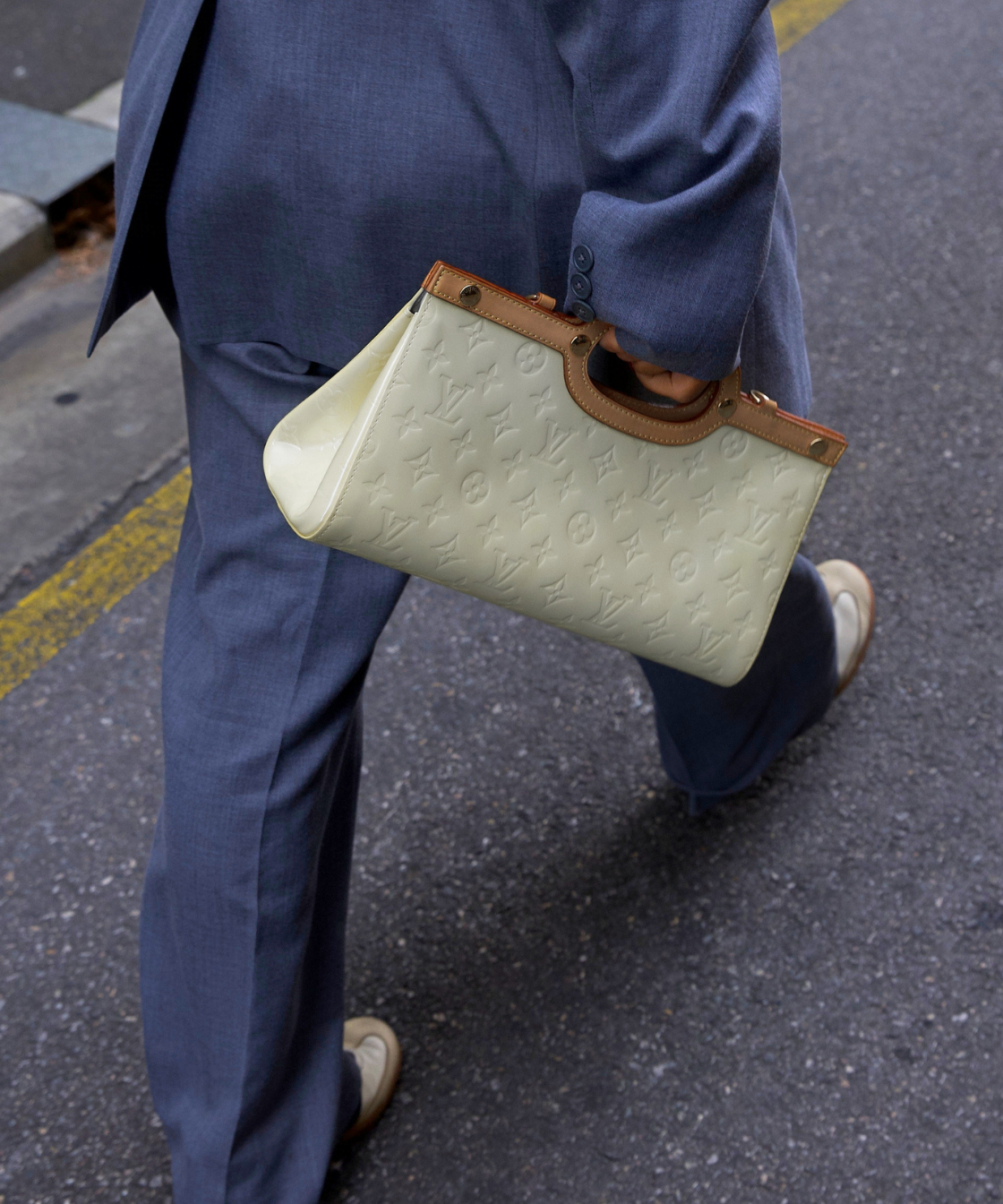 A person in a blue suit walking outdoors while carrying a structured LV coloured embossed leather handbag with brown trim and handle.