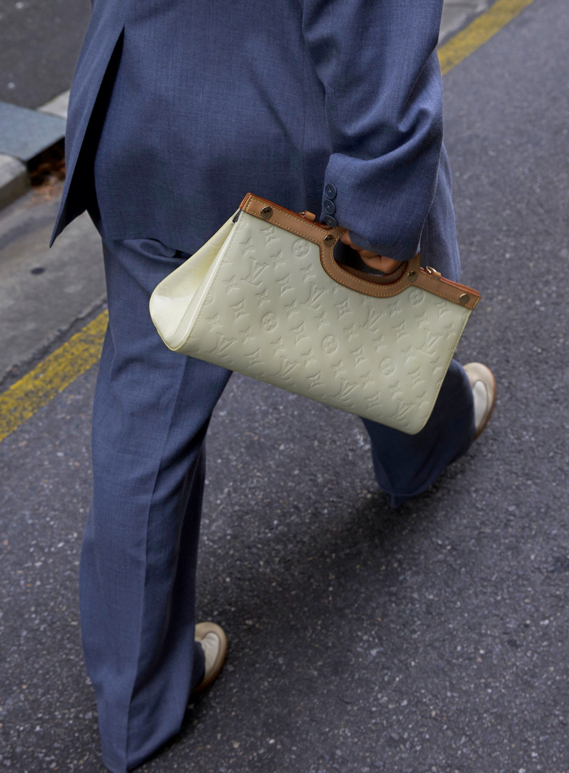 A person in a blue suit walking outdoors while carrying a structured LV coloured embossed leather handbag with brown trim and handle.