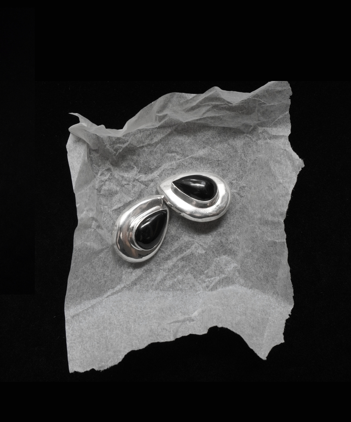 A pair of silver-toned teardrop-shaped earrings with black centers, displayed on a piece of crumpled translucent paper against a solid black background.
