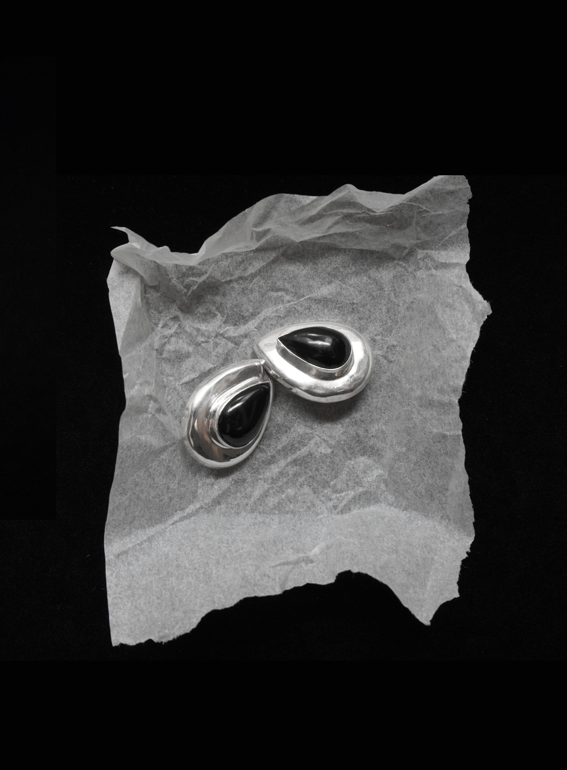 A pair of silver-toned teardrop-shaped earrings with black centers, displayed on a piece of crumpled translucent paper against a solid black background.