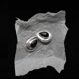 A pair of silver-toned teardrop-shaped earrings with black centers, displayed on a piece of crumpled translucent paper against a solid black background.