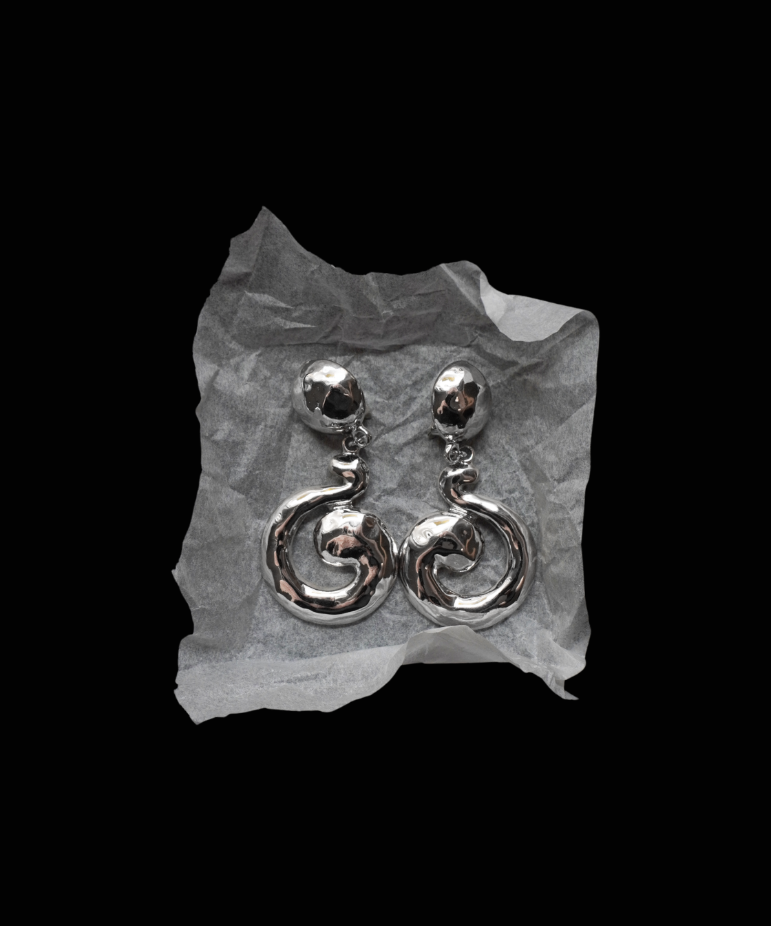 A pair of silver-toned dangle earrings with a textured spiral drop design, displayed on a crumpled piece of white paper against a solid black background.
