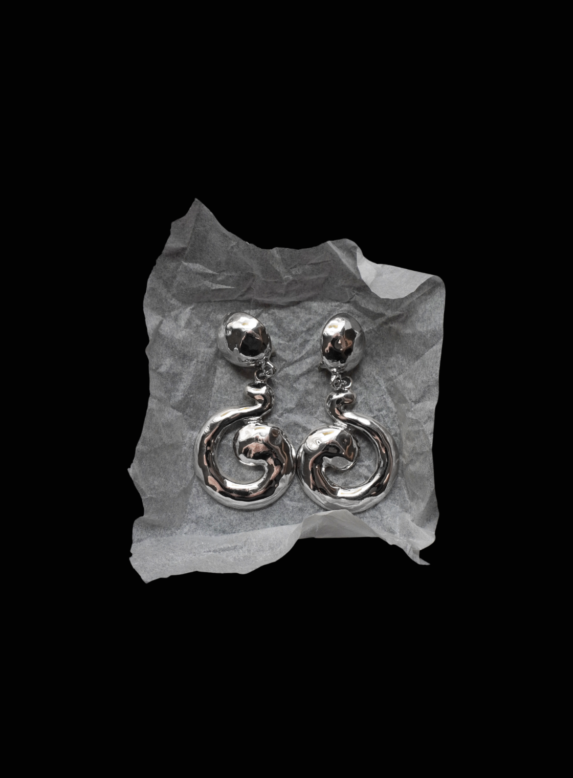 A pair of silver-toned dangle earrings with a textured spiral drop design, displayed on a crumpled piece of white paper against a solid black background.