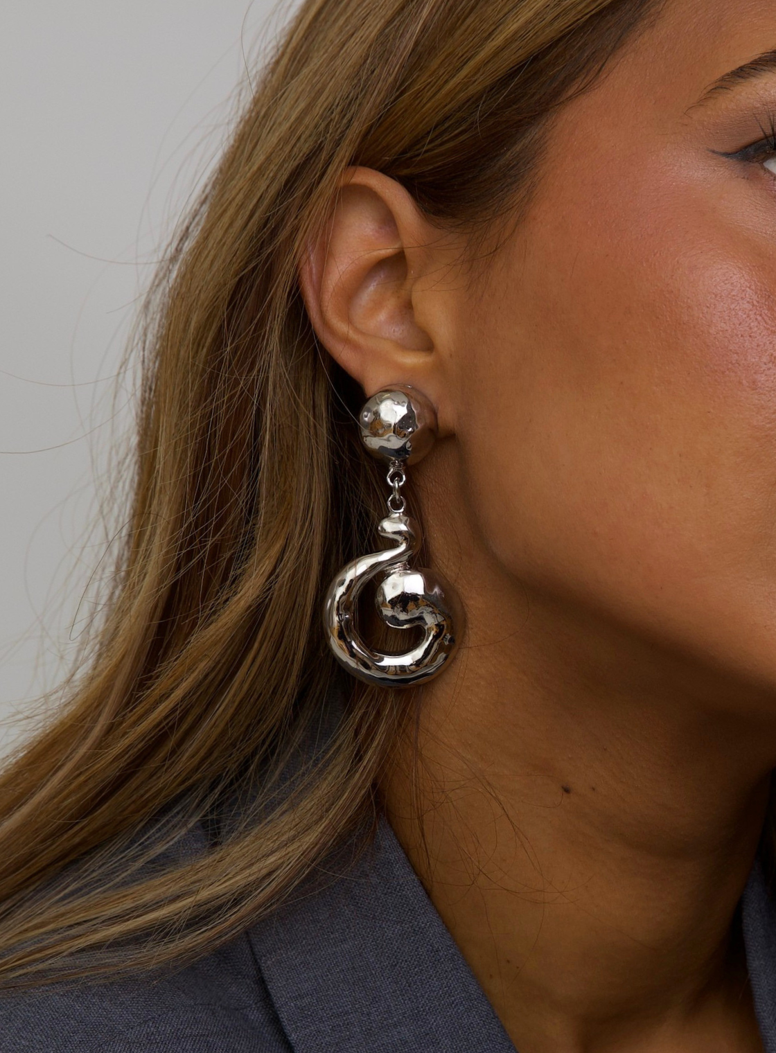 A close-up of a woman's ear wearing a dangling silver earring with a faceted round stud and a large spiral drop charm.