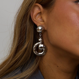 A close-up of a woman's ear wearing a dangling silver earring with a faceted round stud and a large spiral drop charm.