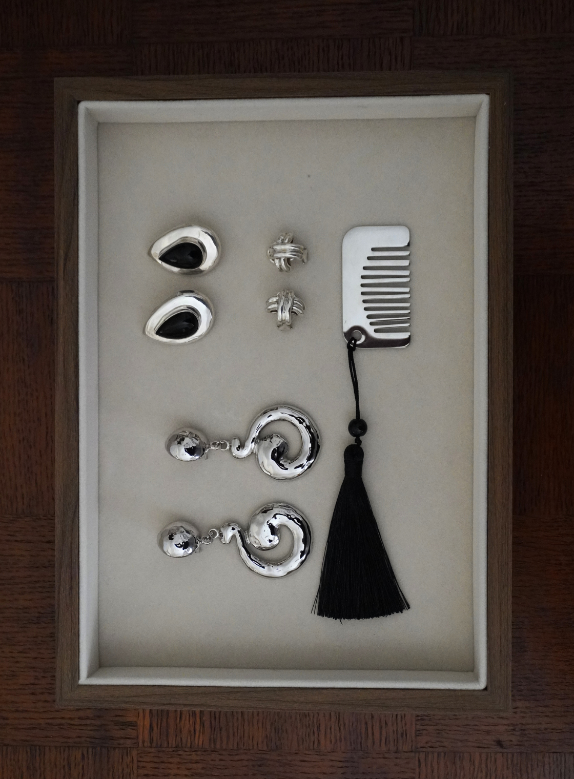 A jewelry gift set in a rectangular presentation box, featuring silver-toned earrings, studs, a comb, and a black tassel, displayed on a beige insert against a dark wood background.