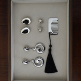A jewelry gift set in a rectangular presentation box, featuring silver-toned earrings, studs, a comb, and a black tassel, displayed on a beige insert against a dark wood background.