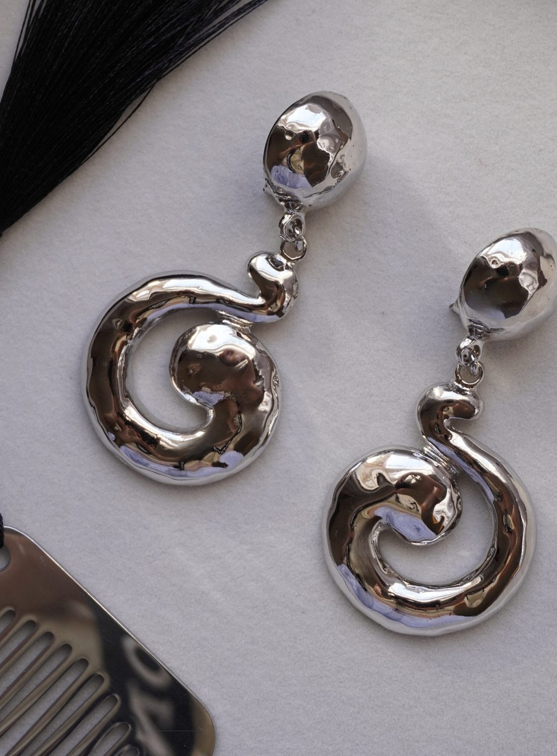 A pair of polished silver drop earrings with a textured, organic spiral dangle design, displayed on a light gray textured surface.