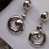 A pair of polished silver drop earrings with a textured, organic spiral dangle design, displayed on a light gray textured surface.