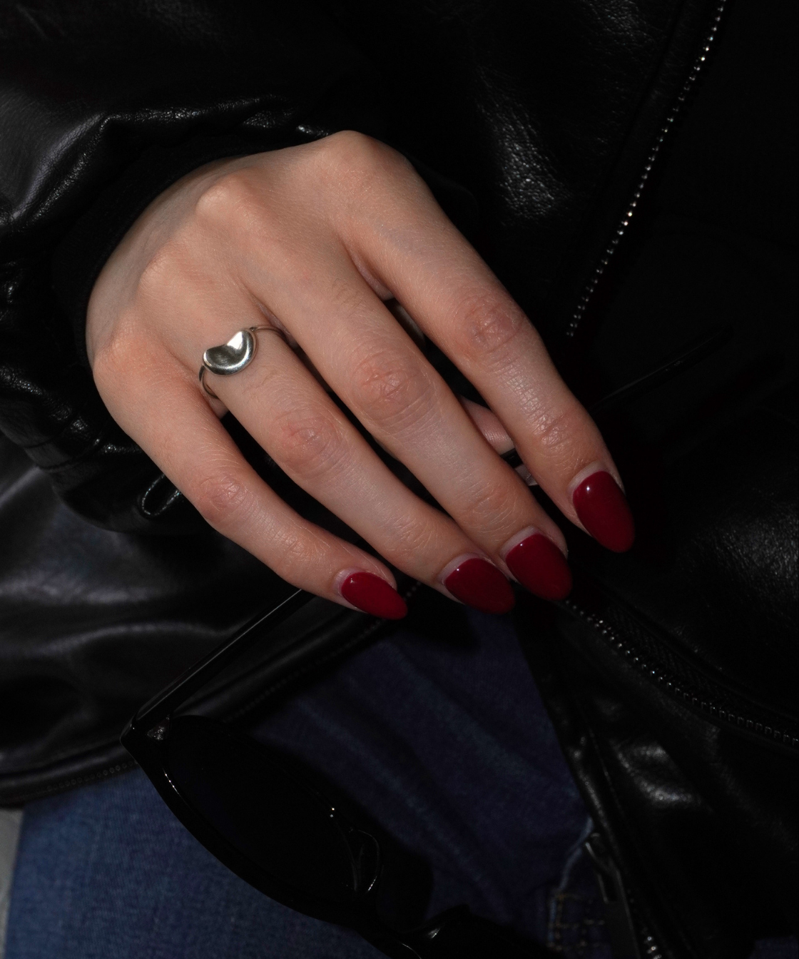 A close-up of a hand wearing a silver ring with a crescent moon design, resting on a black leather jacket. The nails are painted dark red, and blue jeans are visible underneath.