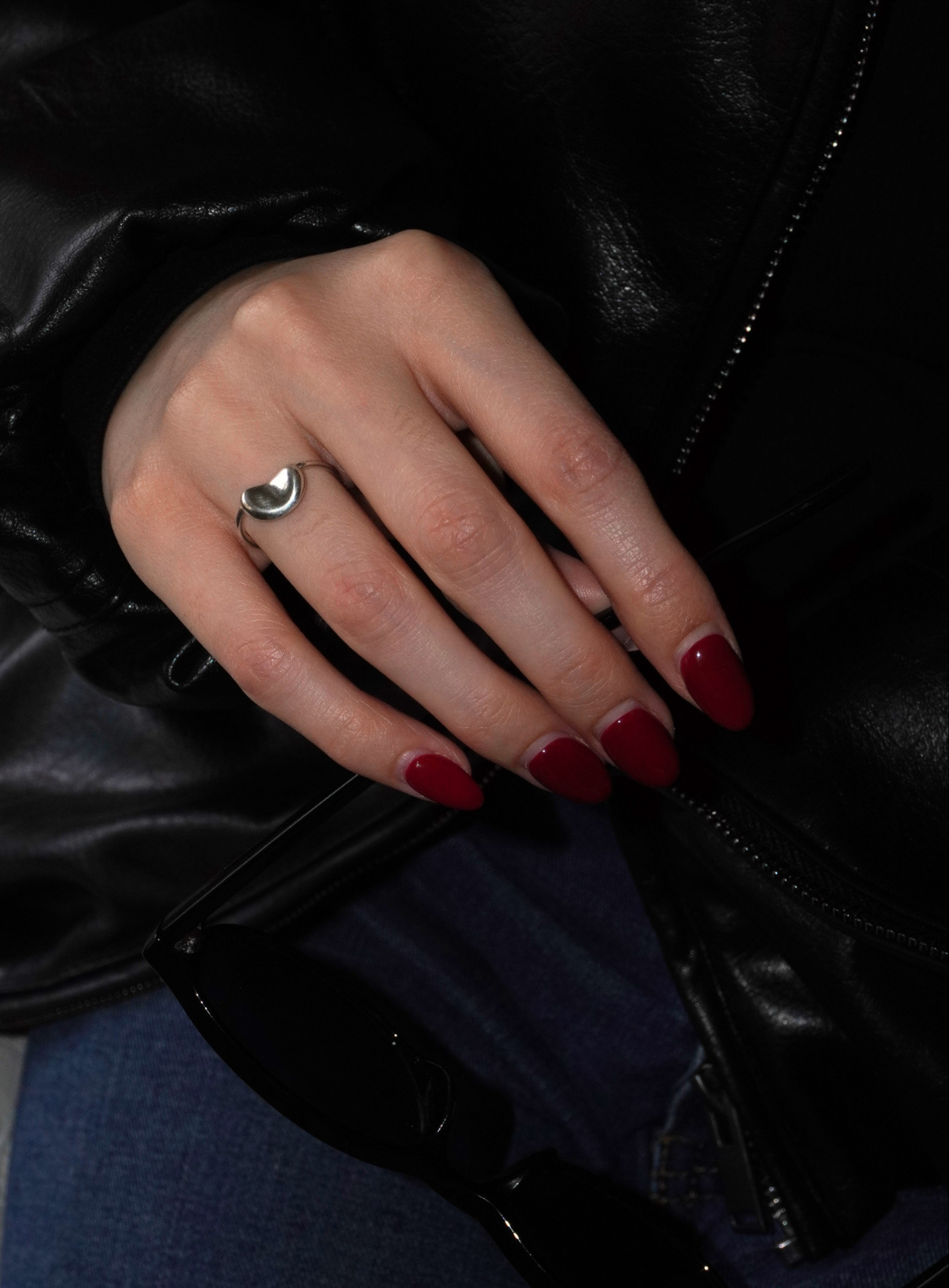 A close-up of a hand wearing a silver ring with a crescent moon design, resting on a black leather jacket. The nails are painted dark red, and blue jeans are visible underneath.