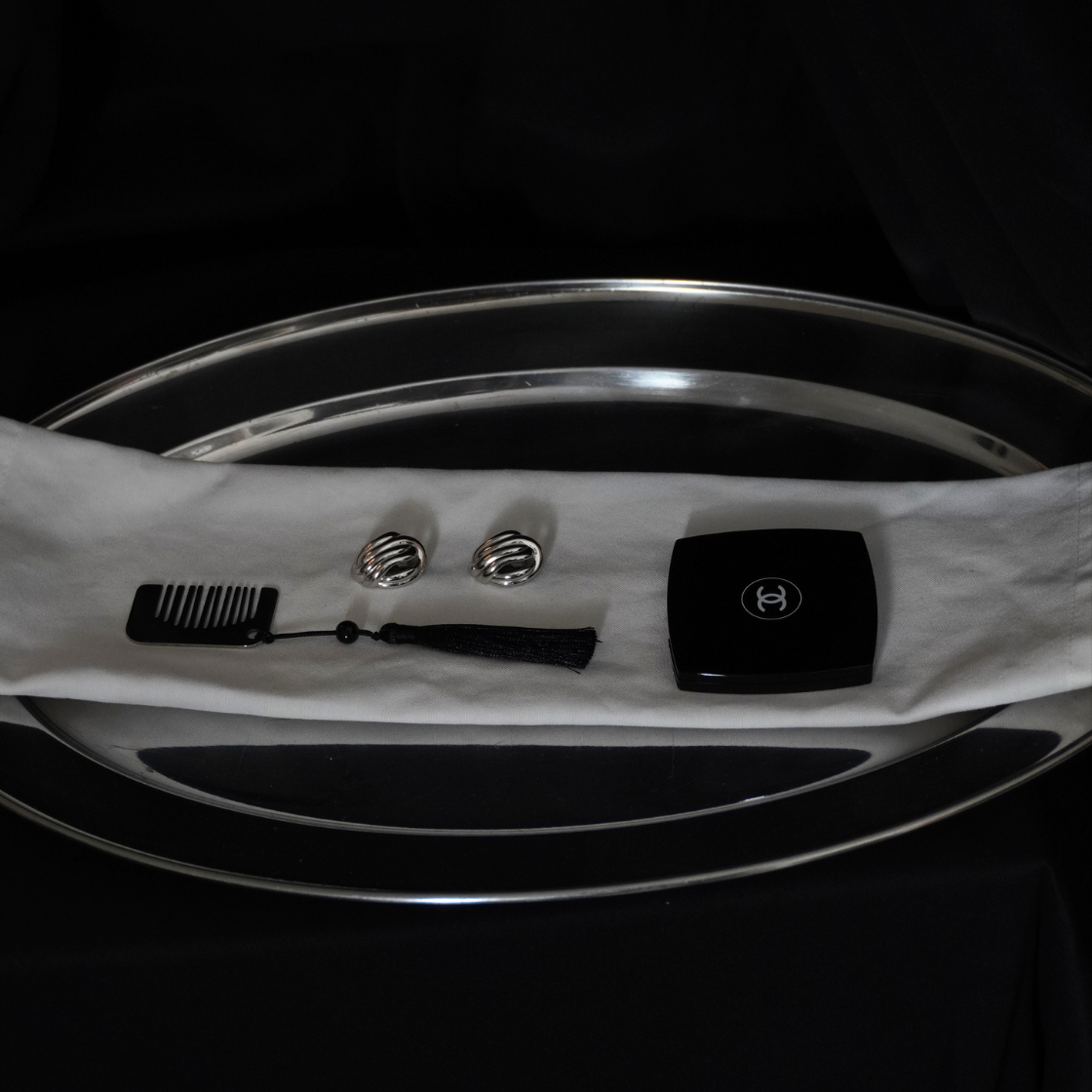 A black and white accessory set displayed on a white cloth over a reflective silver oval tray, featuring a black comb, silver cufflinks, a black necktie, and a black square case with a silver emblem, set against a black background.