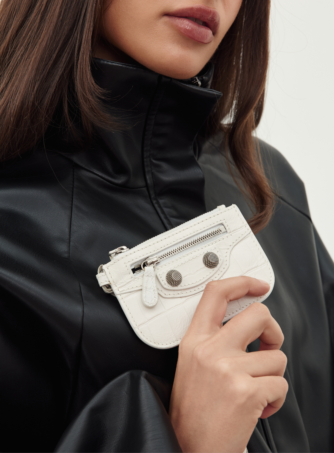 A close-up of a model wearing a black leather jacket and holding a small white leather zip pouch with silver-tone snap buttons and a zipper.