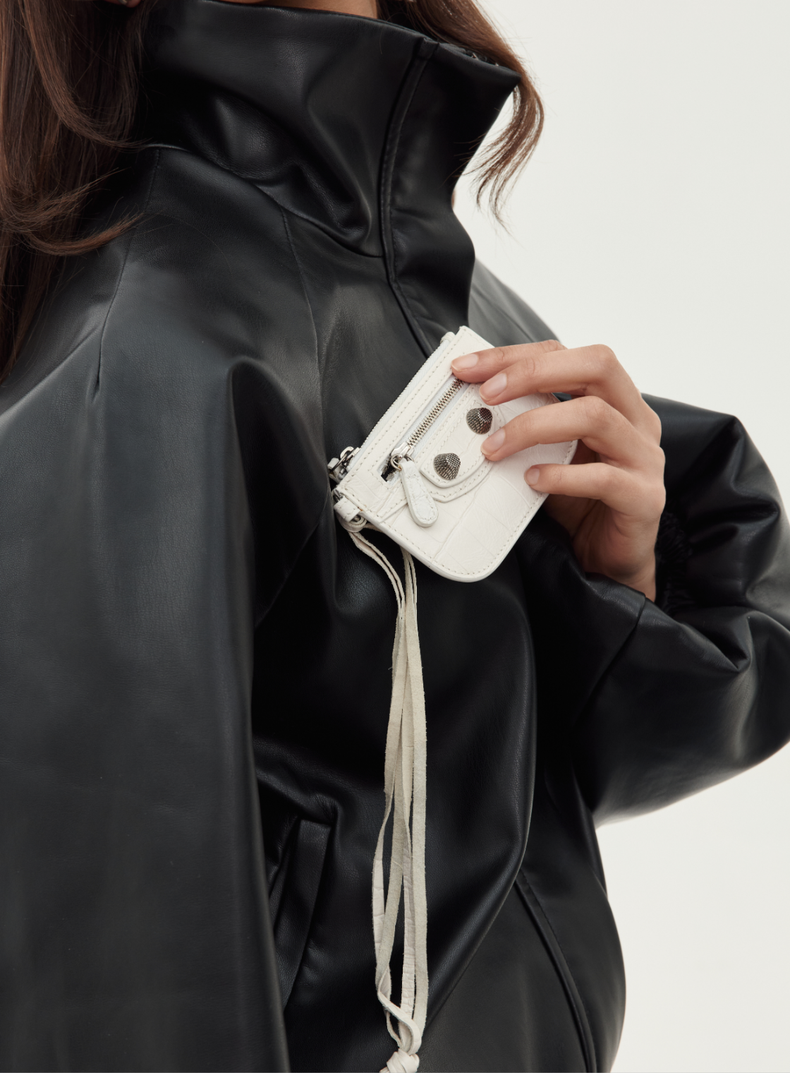 A close-up of a person wearing a black faux-leather jacket and holding a small white leather crossbody bag with silver hardware and a tassel strap.
