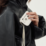 A close-up of a person wearing a black faux-leather jacket and holding a small white leather crossbody bag with silver hardware and a tassel strap.