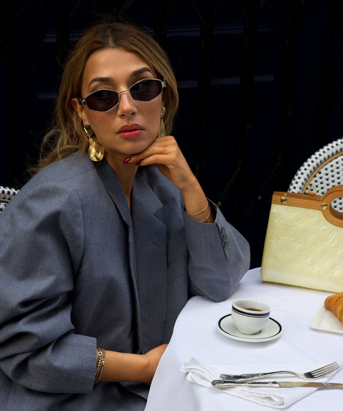 Woman sitting at a table with a cup and saucer, wearing sunglasses and a gray coat.