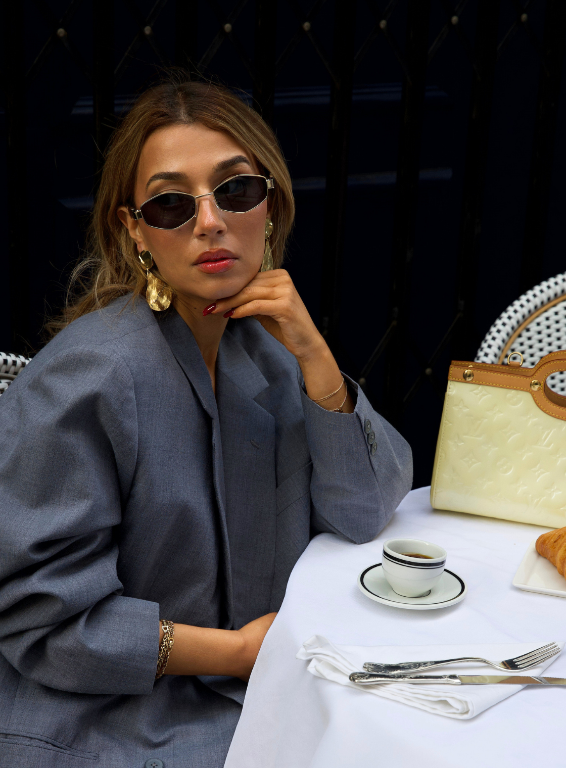 Woman sitting at a table with a cup and saucer, wearing sunglasses and a gray coat.