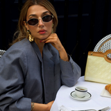 Woman sitting at a table with a cup and saucer, wearing sunglasses and a gray coat.