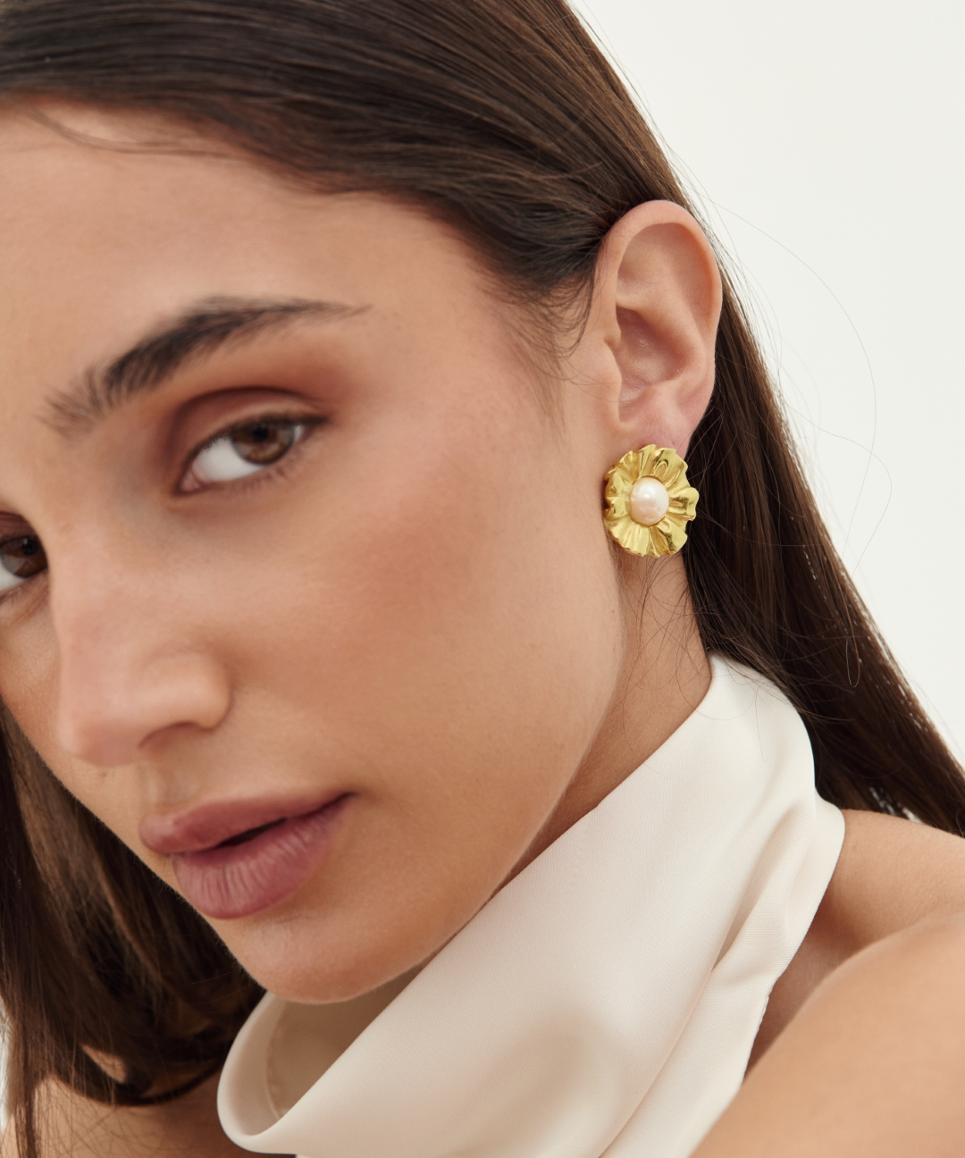 A close-up of a woman wearing a textured gold flower-shaped stud earring with a small white pearl at the center, paired with a cream satin high-neck top against a light off-white background.
