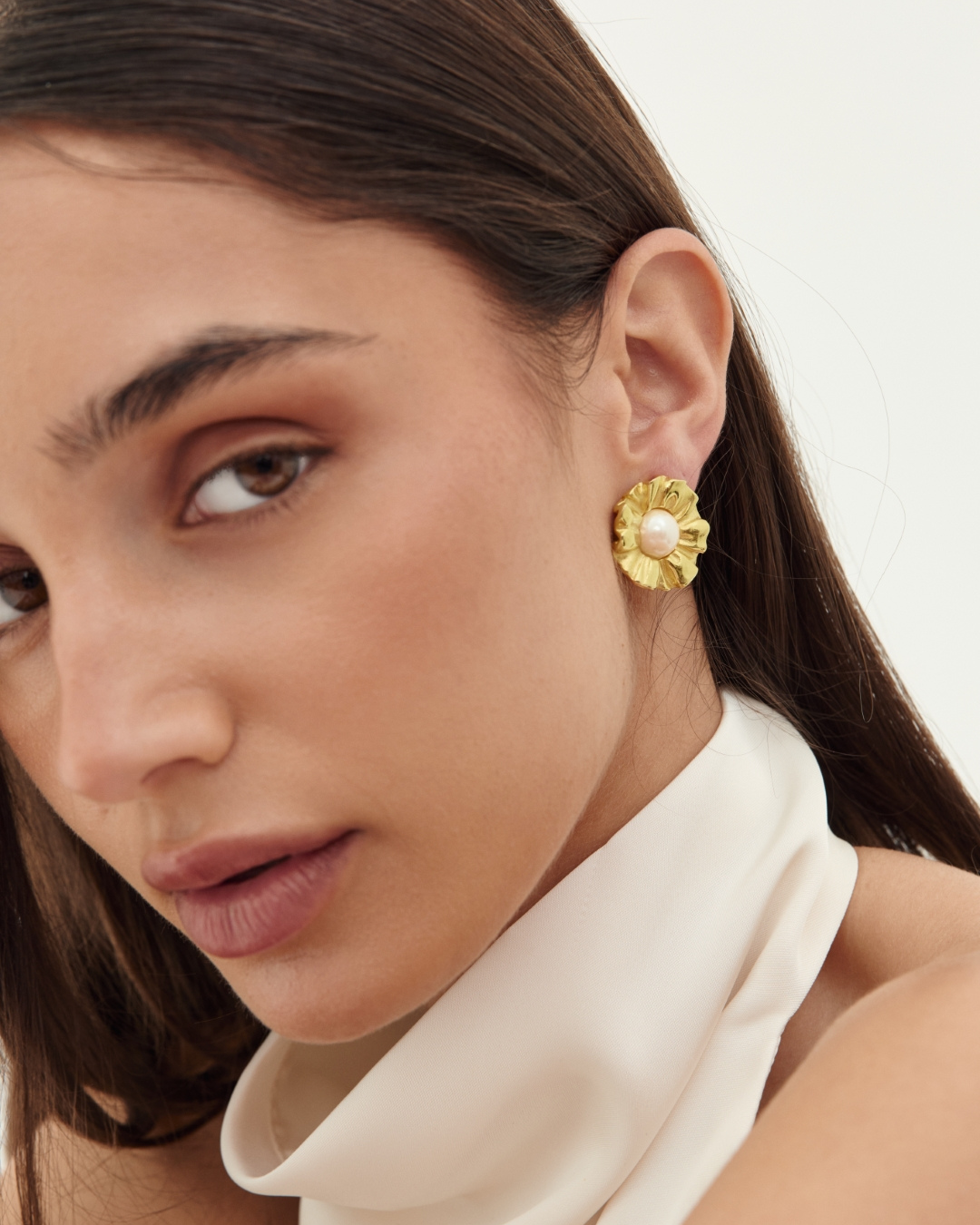 A close-up of a woman wearing a textured gold flower-shaped stud earring with a small white pearl at the center, paired with a cream satin high-neck top against a light off-white background.