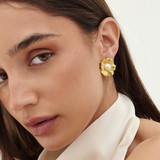 A close-up of a woman wearing a textured gold flower-shaped stud earring with a small white pearl at the center, paired with a cream satin high-neck top against a light off-white background.